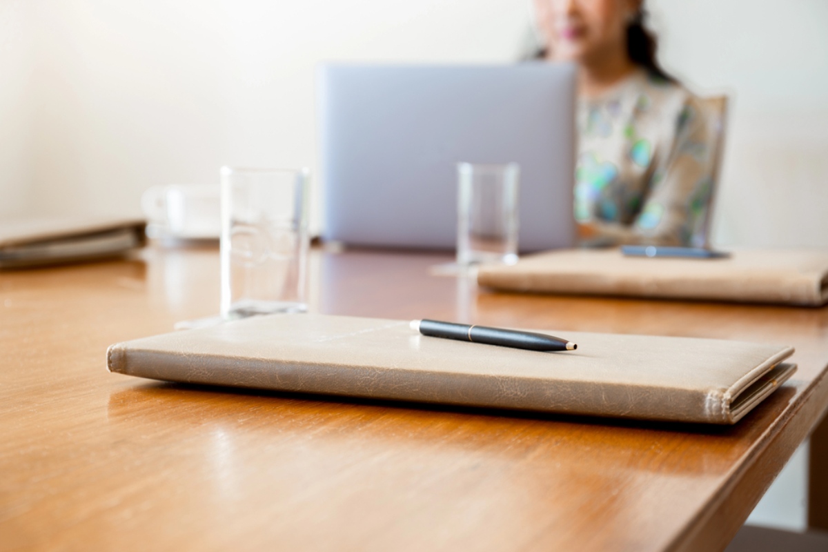 Tavolo d’ufficio con collega al computer e agenda con penna in primo piano.